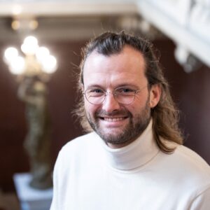 Portrait of Maxi Guennewig, CERGE-EI faculty member, smiling at the camera in a white turtleneck sweater and round glasses, with a softly blurred interior in the background.